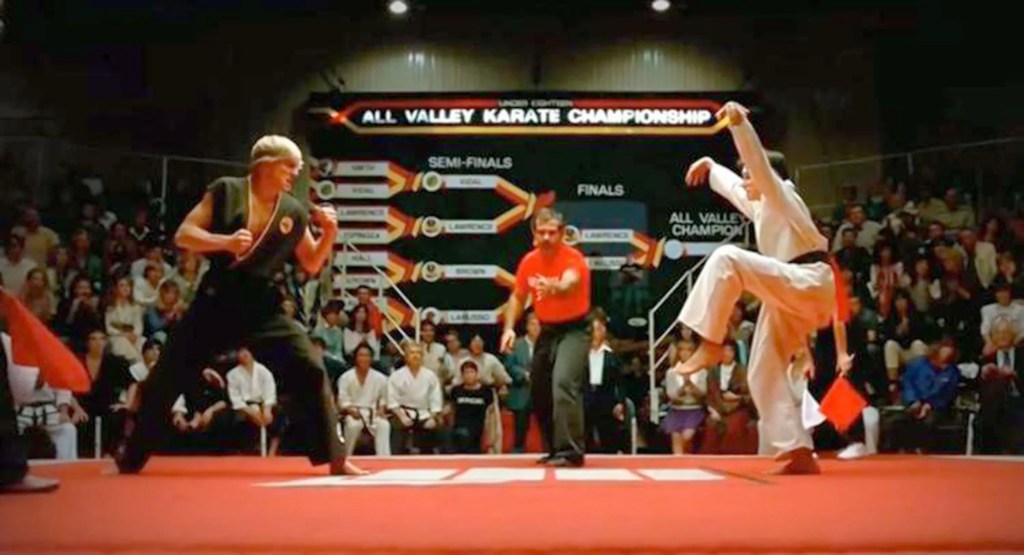 Dennis's alt text: Ralph Macchio balances on one foot with his hands in the air as the dastardly Cobra Kai Johnny prepares his attack. My addition: they are at a competition with a red-shirted ref between them and a shocking amount of people on the bleachers behind them