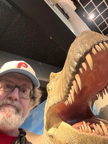 Dennis is wearing a Phillies cap and is next to a dinosaur at the Natural History Museum.