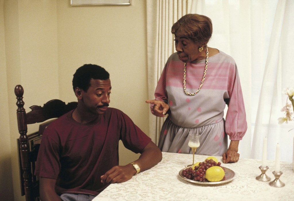 A mustached Black man in a maroon t-shirt sits at an elegant table in front of a plate of fruit while an older Black woman points her finger at him in seeming disapproval.