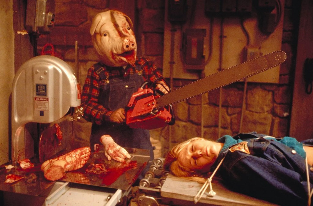 A person in a pig mask? with a pig head? wears flannel and denim overalls while wielding a chainsaw, a woman is tied to a coveyer belt. There are bloody hands and feet by her head.