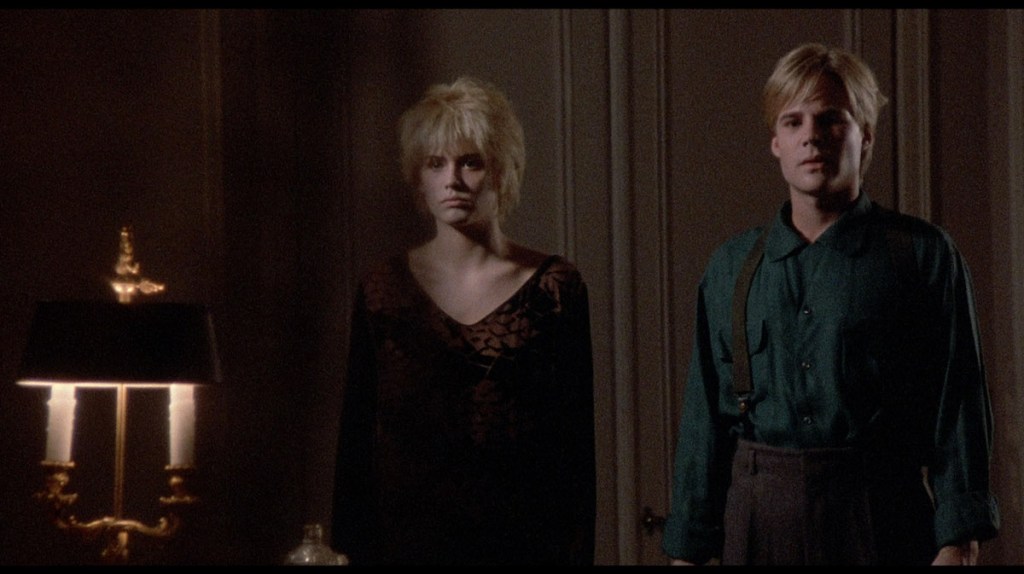 Two pasty blond "teens" (one in a truly terrible wig) stand side by side in a dim room